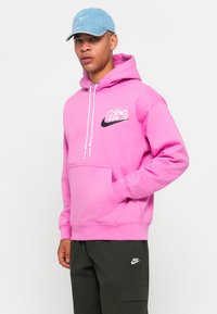 Felpa rosa con coulisse bianca e tasca frontale, con logo Nike nero. Abbinata a pantaloni cargo verde scuro.