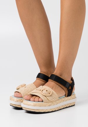 Sandales tressées beiges avec une plateforme, présentant un design texturé et un accent rond proéminent, et une sangle noire réglable autour de la cheville.