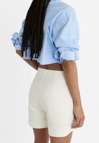 Camicetta a righe blu chiaro, corta, con maniche arrotolate, abbinata a pantaloncini crema. Tessuto morbido, vestibilità su misura e dettaglio di cuciture minimale.