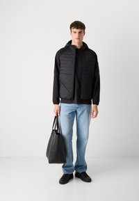 Schwarze Pufferjacke mit Kapuze und strukturiertem Vorderteil, kombiniert mit einem schwarzen Rundhals-Shirt, hellblauen Jeans und einer schwarzen Leder-Tote.