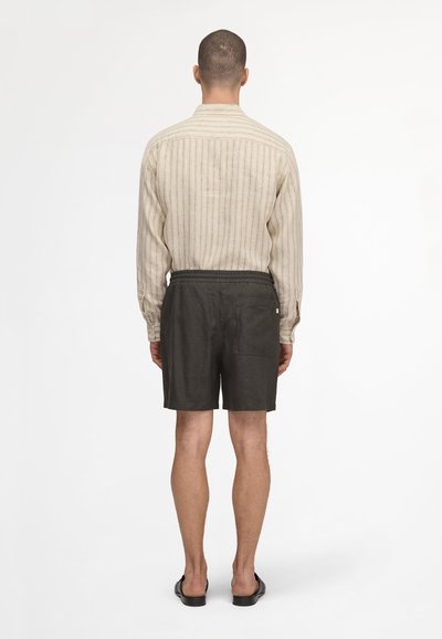 Homme debout, de dos, portant une chemise beige rayée à manches longues, un short noir avec une poche arrière, et des sandales noires sur fond blanc.