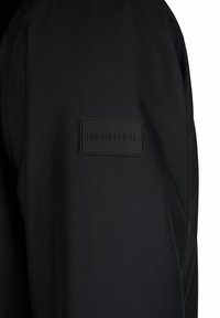 Tissu noir texturé avec un écusson rectangulaire noir portant l'inscription « HECHTER » cousu sur la manche d'un vêtement.