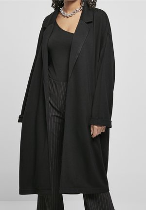 Persoon draagt een loszittende zwarte jas over een zwart tanktopje en zwarte broek met fijne strepen, geaccentueerd met een dikke zilveren ketting.