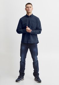 Marineblå skjorte med knapper og frontlomme, kombinert med mørk denimjeans og svarte treningssko, stående mot en lys bakgrunn.