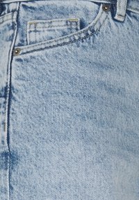 Světle modré džíny z denimu s viditelnou přední kapsou, žlutým vrchním prošitím a mosazným kováním. Materiál má měkkou strukturu s mírným vybledlým efektem.