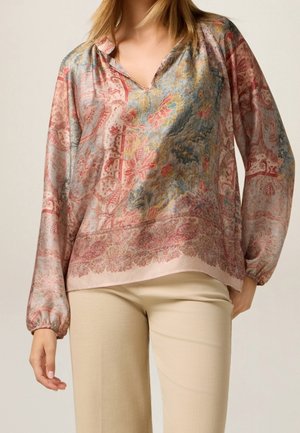 Kobieta ubrana w bluzkę z długim rękawem w kwiatowy wzór z dekoltem w serek oraz beżowe spodnie, stojąca na neutralnym tle.