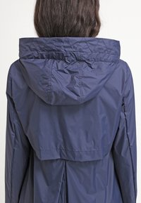 Veste imperméable bleu marine avec capuche matelassée, dotée d'un cordon de serrage au niveau du col et d'une coupe décontractée, fabriquée à partir d'un matériau lisse et léger.