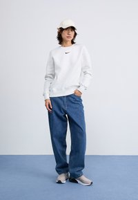 Lichtgrijze sweatshirt met een zwart logo, gecombineerd met losse blauwe denim jeans. Een beige pet en grijze sneakers maken de outfit compleet.