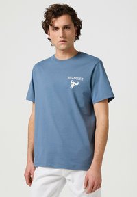 Camiseta de algodón azul con cuello redondo, mangas cortas y un logotipo blanco de "WRANGLER" con un diseño gráfico en la parte superior izquierda.