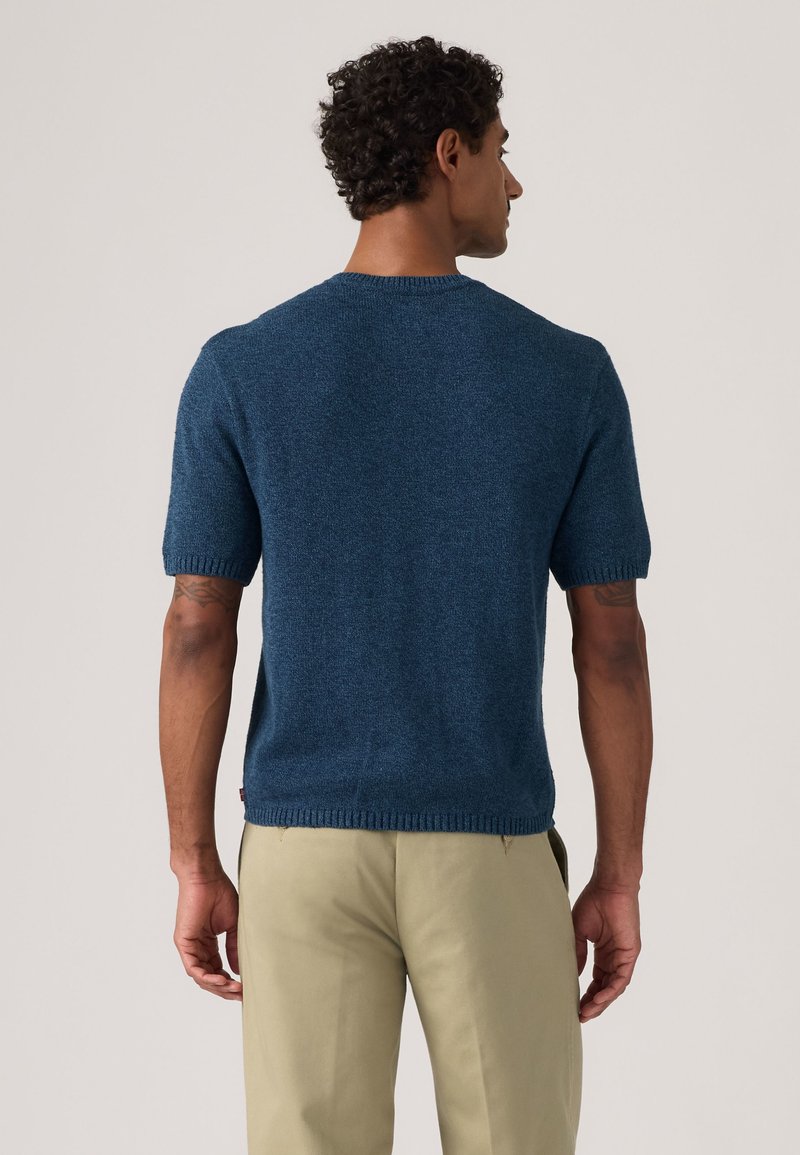 Mann mit lockigem Haar, der abgewandt ist und einen kurzärmligen blauen Strickpullover sowie beige Hosen vor einem schlichten hellen Hintergrund trägt.