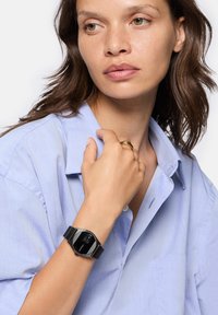 Schwarze Uhr mit rundem Ziffernblatt und digitaler Anzeige an einem schwarzen Armband, getragen an einer Hand gegen ein hellblaues Hemd. Glatte Textur, minimalistisches Design.