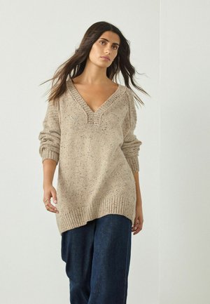 Beige gestrickter Pullover mit tiefem V-Ausschnitt, geripptem Saum und rollierten Ärmeln, der ein strukturiertes Muster und subtile Sprenkel aufweist.