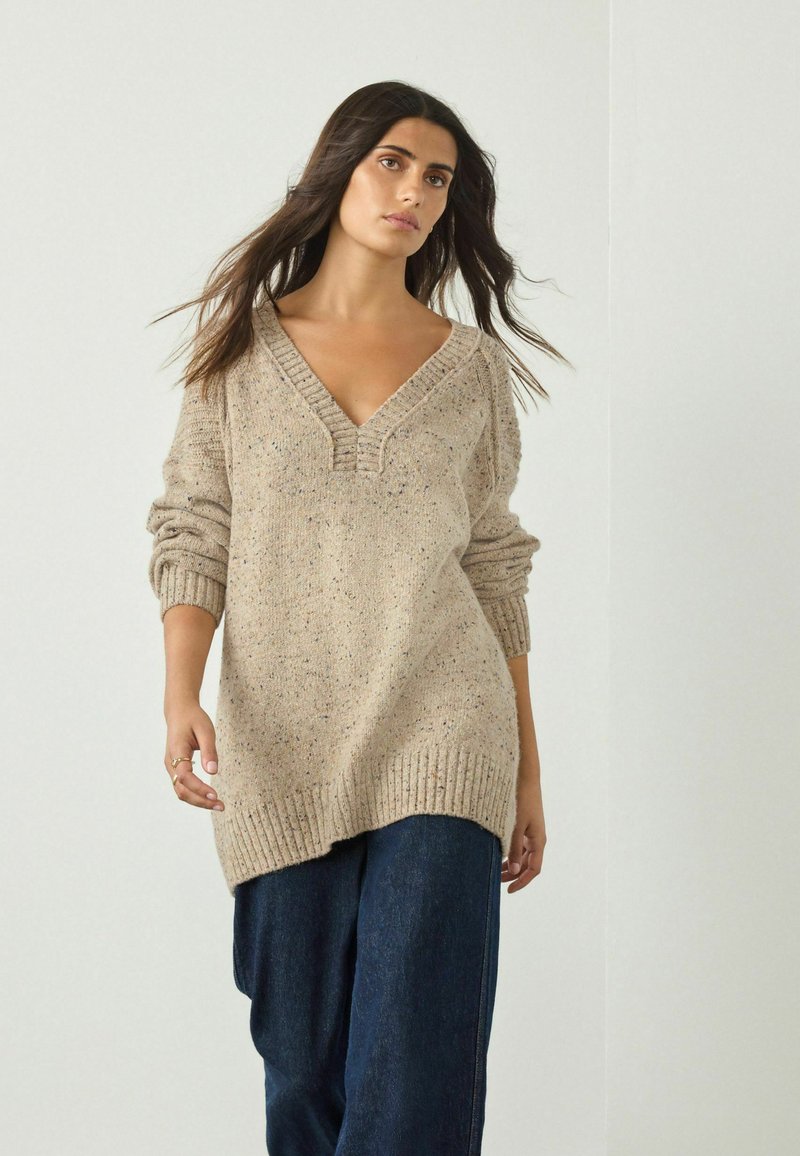 Beige strikket sweater med dyb V-hals, ribbet kant og opskruede ærmer, der har et struktureret mønster og subtile prikker gennem hele.