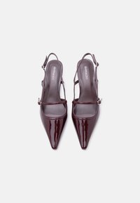 Paire d'escarpins slingback brillants rouge foncé à bout pointu avec fines brides et boucles argentées sur fond blanc.