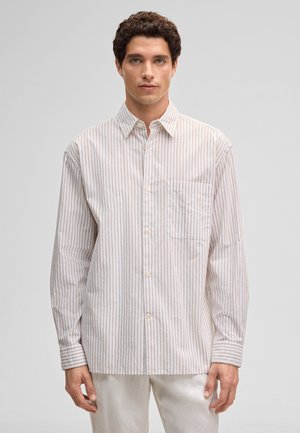 Uomo con capelli ricci castani che indossa una camicia a righe verticali beige e bianche con bottoni e pantaloni bianchi, in piedi davanti a uno sfondo semplice.