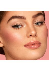 Close-up van een gezicht met een egale huid, gedefinieerde wenkbrauwen en volle lippen met een lichte glans. Ogen met eyeliner op een roze achtergrond.