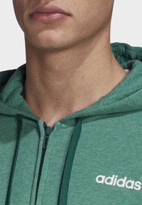 Sudadera verde con cremallera fabricada en tejido suave, con una cremallera verde oscuro, cordones y un logo blanco en el lado izquierdo del pecho.