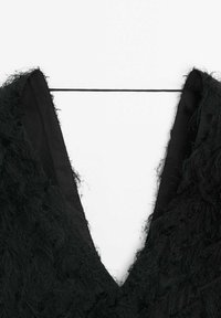 Tissu noir texturé avec un décolleté en V profond maintenu par une fine bretelle traversant la partie supérieure de la poitrine sur fond blanc.