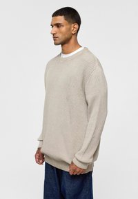 Beige gebreide trui met ribbeltextuur, ronde hals, losse pasvorm en lange mouwen. In combinatie met donkerblauwe wijde jeans.