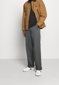 Bruine canvasjas met twee voorzakken, zwart t-shirt, grijze broek met brede pijpen en witte Nike sneakers met zwarte accenten.