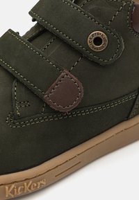 Zapato de ante verde oscuro con dos correas de gancho y bucle, acentos de cuero marrón, suela de goma beige y logotipo en relieve en el lado.