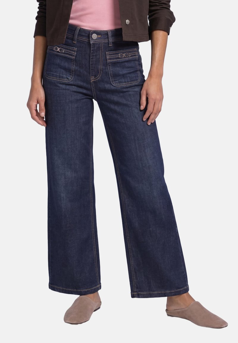 Femme portant un jean large bleu foncé avec poches appliquées à l'avant, un haut rose, une veste marron foncé et des chaussures taupe à enfiler.
