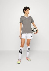 Nő szürke rövid ujjú pólóban, fehér sportnadrágban, fekete-fehér focilabdát tartva, fehér térdzokniban és színes cipőkben.