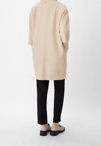 Beige ylipitkä vaate, jossa on lyhyet hihat, sivuhalkiot ja ribbineulos, yhdistetty mustiin housuihin ja vaaleanharmaisiin kenkiin.