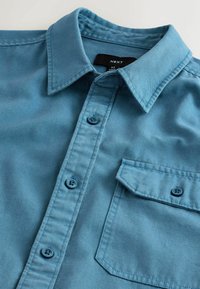 Lichtblauwe overhemdbloes met een kraag, twee voorzakken en blauwe knopen. Gemaakt van textuurstof, met een eenvoudige, rechttoe-rechtaan ontwerp.