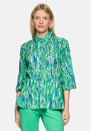 Woman wearing a blue, green, and white patterned button-up blouse with 3/4 sleeves and solid green pants, standing against a plain background.