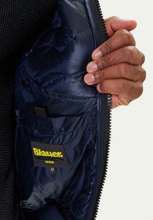 Intérieur d'une doudoune matelassée bleu marine avec une texture matelassée et une étiquette portant "Blauer" en jaune. Une poche à l'intérieur, fermeture éclair visible le long du bord.