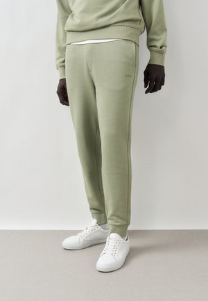 Man wearing light green jogger pants, white sneakers, and matching sweatshirt standing against white background.