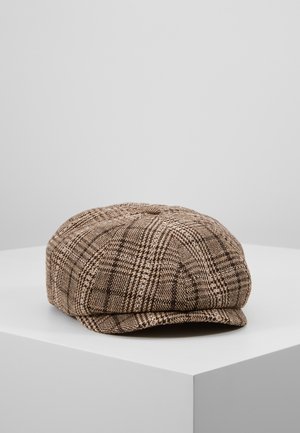 Casquette plate à carreaux marron et beige reposant sur un socle blanc sur fond gris clair.