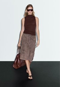 Haut sans manches marron avec col roulé, jupe en suède beige à mi-mollet, sandales ouvertes marron et un grand sac en cuir marron. Design simple.