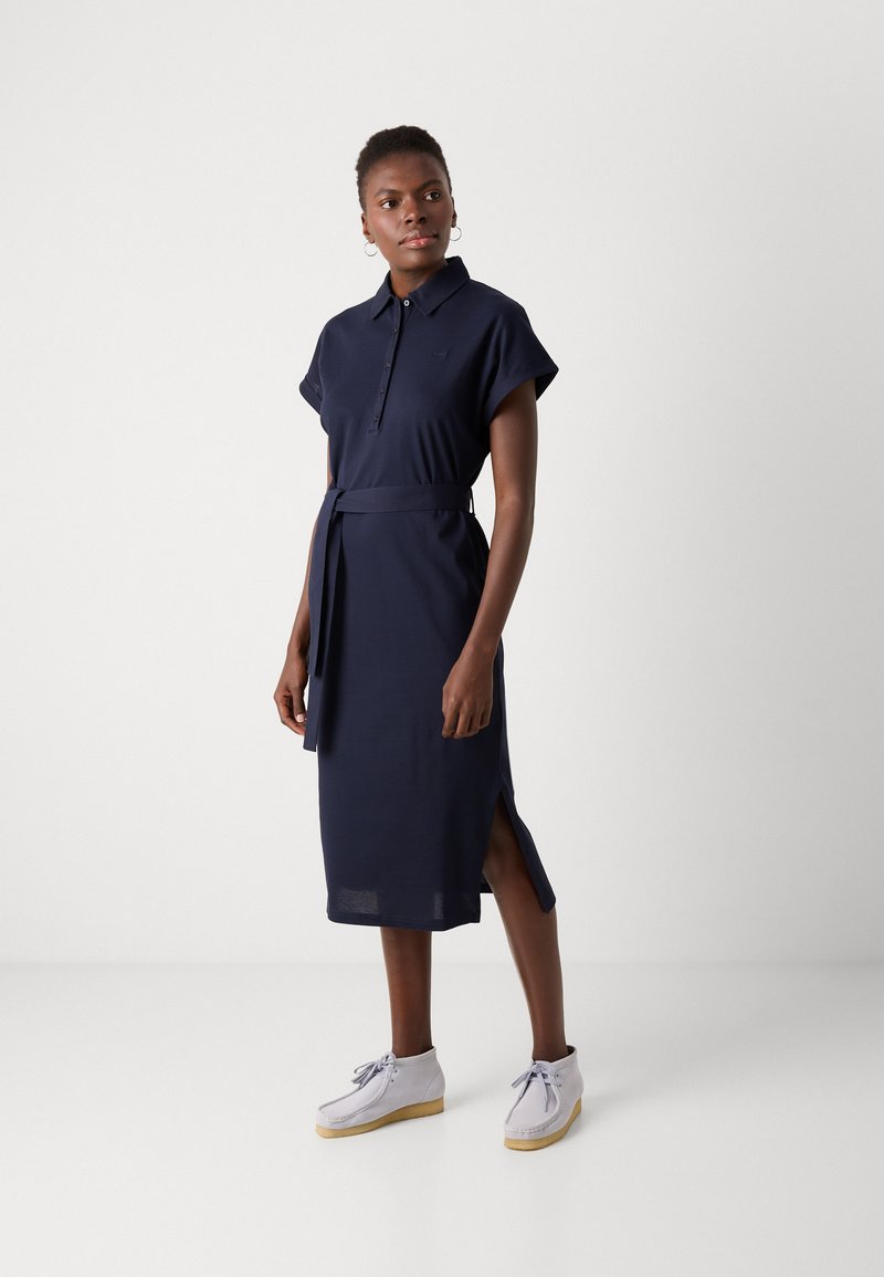 Granatowa sukienka polo z krótkimi, podwiniętymi rękawami, z zapięciem na guziki i związanym w pasie. Posiada boczne rozcięcia dla swobody ruchów.