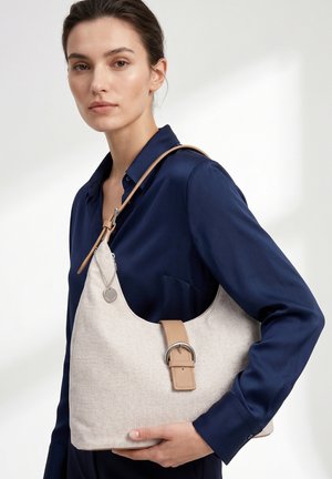 Vrouw in marineblauw overhemd houdt een beige gestructureerde schoudertas met leren band en metalen gesp vast tegen een effen witte achtergrond.