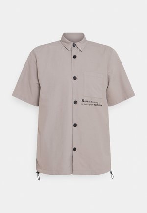 Chemise beige à manches courtes avec poche poitrine et texte « AYEZ le COURAGE de partager vos SENTIMENTS » sur le côté de la poche.