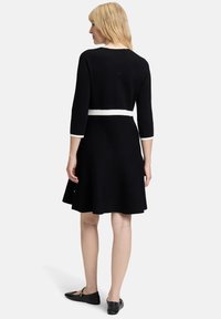 Robe noire avec des manches trois-quarts, détail de ceinture blanche, jupe évasée et design simple. Associée à des ballerines noires.
