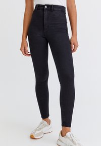 Jeans de mezclilla negros de cintura alta con una forma ajustada, que presentan un cierre de botón y bolsillos laterales, combinados con zapatillas deportivas blancas.