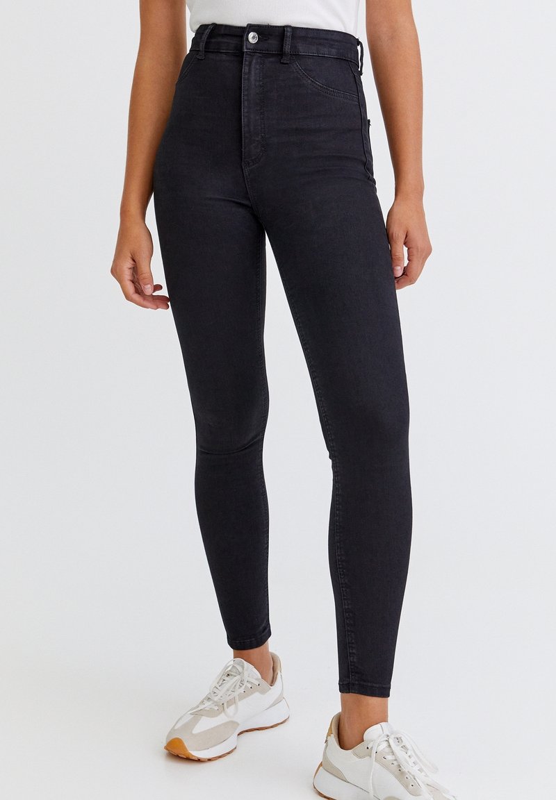 Jeans de mezclilla negros de cintura alta con una forma ajustada, que presentan un cierre de botón y bolsillos laterales, combinados con zapatillas deportivas blancas.