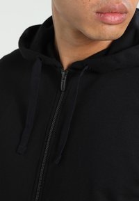 Sweat à capuche zippé noir avec cordon de serrage, tissu doux et design décontracté. Vue rapprochée du col et de la zone de la bouche.