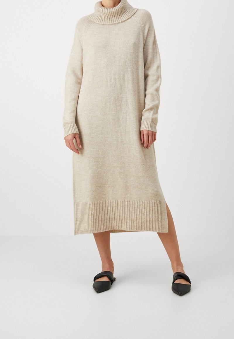 Femme portant une robe pull longue beige en tricot à col roulé, associée à des ballerines noires pointues sur fond blanc.