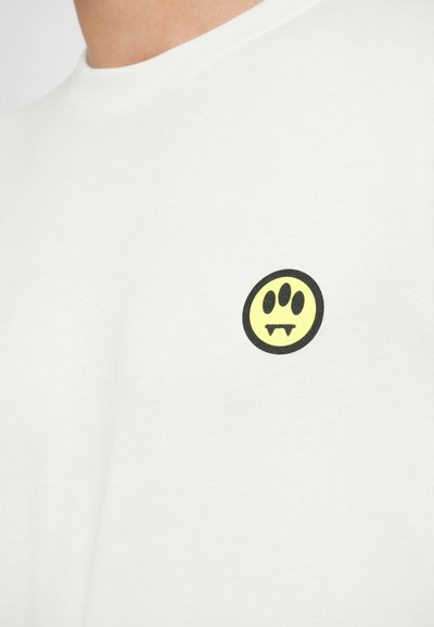 Camiseta blanca de algodón con un pequeño gráfico redondo negro que presenta una cara sonriente amarilla con colmillos, ubicado en el lado izquierdo del pecho.