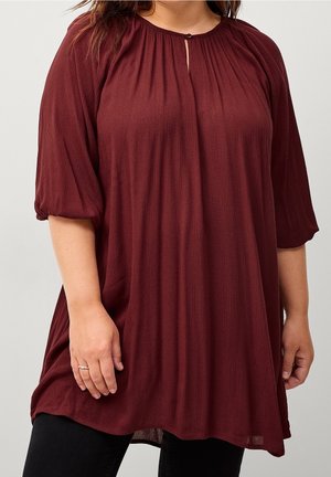 Nainen pukeutuneena väljään, viininpunaiseen puseroon, jossa on kolme neljänneksen mittaiset liehuhihat ja pieni etuosan nappikiinnitteinen avainreikäyksityiskohta.