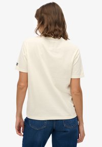 Bawełniana koszulka w kremowym kolorze, z krótkimi rękawami, okrągłym dekoltem i małym logo na lewym rękawie. Materiał wydaje się miękki i gładki.