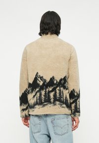 Beige Fleecejacke mit einem gebirgigen Landschaftsdesign in dunklen Tönen. Sie verfügt über einen hohen Kragen und eine entspannte Passform, kombiniert mit hellblauen Jeans.