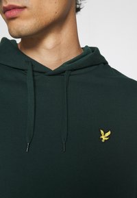 Hoodie verde scuro realizzato in tessuto liscio, con un logo dell'aquila ricamato in giallo sul petto e cordini sul cappuccio.