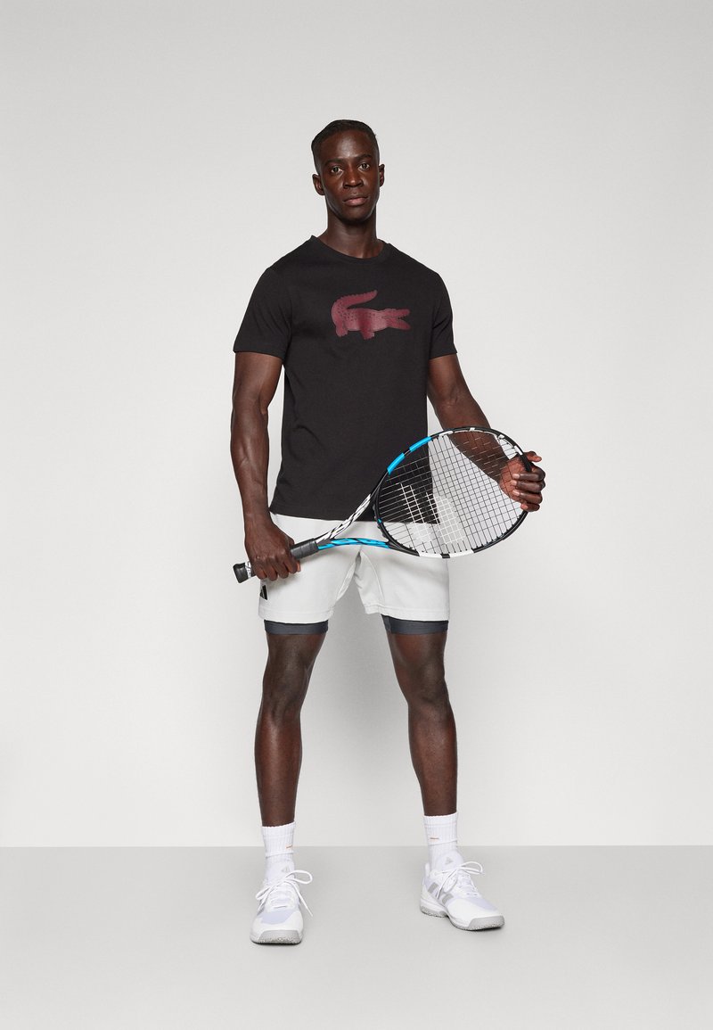 T-shirt noire avec un logo crocodile bourgogne, shorts blancs avec des accents foncés, et chaussures de tennis blanches. Tenant une raquette de tennis.