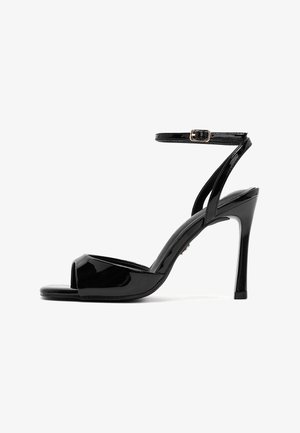 Sandalo con tacco alto in vernice nera, punta aperta, cinturino alla caviglia e tacco a spillo sottile su sfondo bianco.