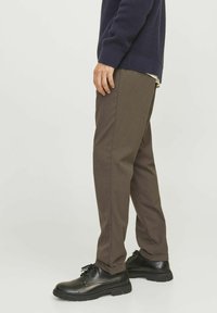Braune maßgeschneiderte Hosen aus einem glatten Stoff, kombiniert mit schwarzen Schnürschuhen. Das Outfit umfasst einen strukturierten marineblauen Pullover.